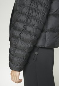 Chaqueta de plumas negra con secciones acolchadas y cosidas, un cuello alto y puños elastizados. Combinada con pantalones negros ajustados y detalle de cordón.