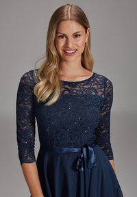 Laivastonsininen mekko, jossa on pitsinen bodice kukkakuviolla, kolmannelta neljännekseltä hihat ja satiininen hame, jossa on solmittava vyötärönauha.