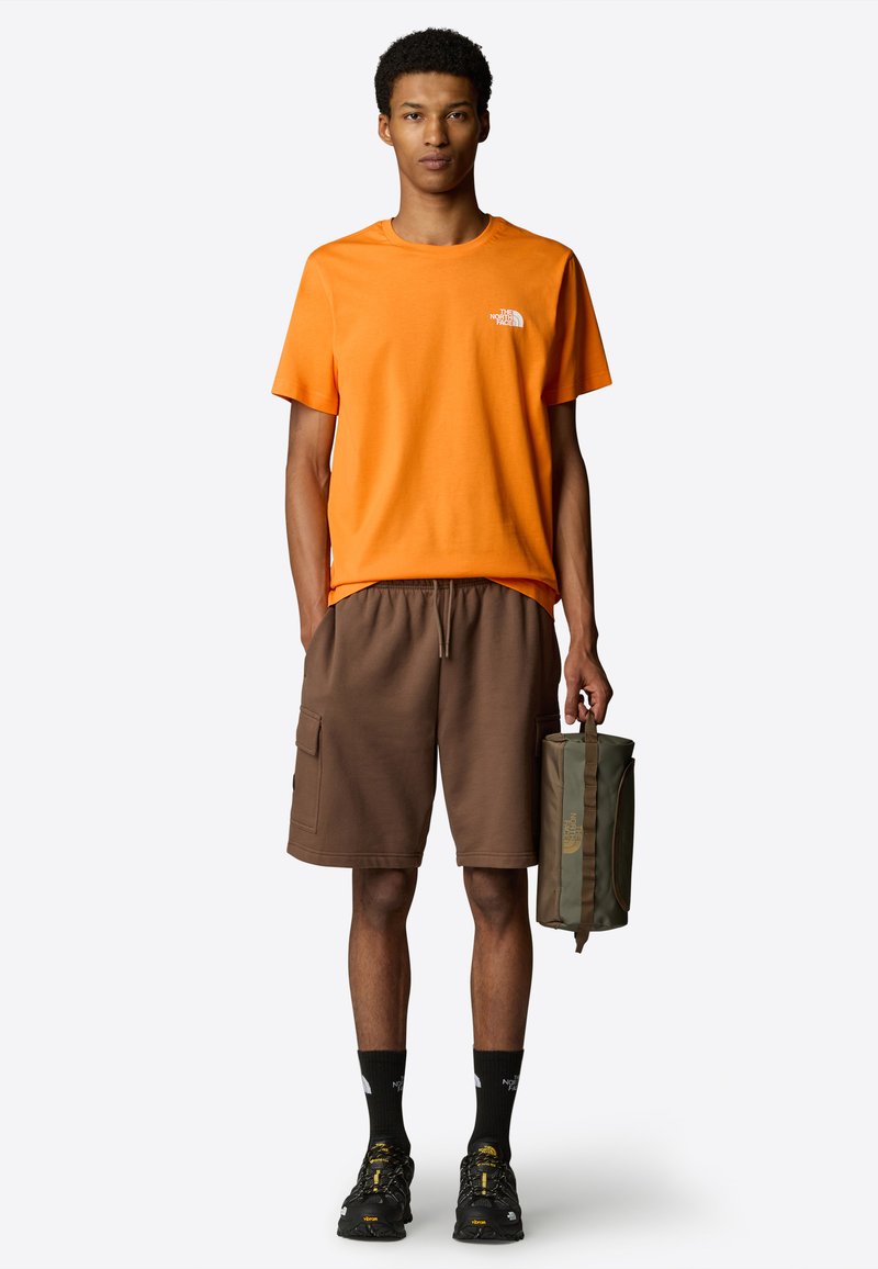 Orange t-shirt med kort ärm, bruna cargo-shorts med sidofickor, svarta sneakers och grön väska. Enkelt, casual design.