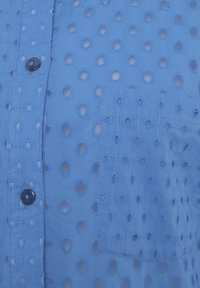 Camicia blu con bottoni, caratterizzata da modelli circolari ritagliati, presenta una tasca a toppa e dettagli di cucitura testurizzati. Tessuto morbido con un aspetto leggero.
