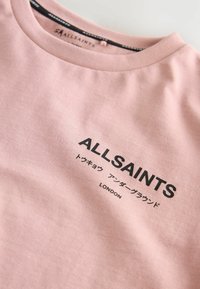Rožinė medvilninė marškinė su apvaliu kaklu. Ant jų yra juodu tekstu užrašas "ALLSAINTS" ir papildomas tekstas japonų kalba, o po juo - "LONDON".