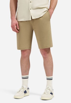 Man draagt beige knielange shorts, witte sneakers met marineblauwe accenten en witte sokken met zwarte strepen, staand tegen een witte achtergrond.