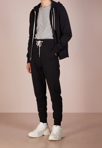 Schwarze Kapuzenjacke mit weißen Kordeln über einem grauen T-Shirt, kombiniert mit schwarzen Jogginghosen und weißen klobigen Sneakern. Glatte Stoffstruktur.