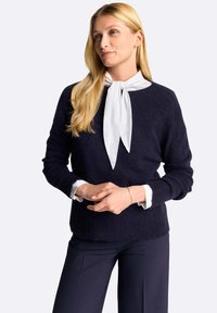 Maglione lavorato a maglia di colore blu navy con maniche lunghe e finitura testurizzata, indossato sopra una camicia bianca con colletto e dettaglio di fiocco legato al collo.