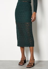 Falda lápiz de encaje verde esmeralda con dobladillo de onda, que presenta un diseño ajustado que llega a la mitad de la pantorrilla. Combinada con sandalias negras de tiras.