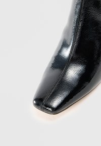 Bota de tobillo de charol negro con punta afilada y costuras visibles. Textura suave con un acabado brillante.