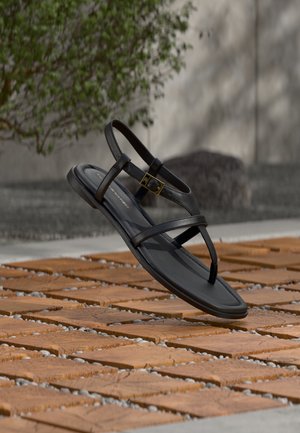 Sandalia plana negra con tiras y hebilla dorada flotando sobre un suelo con patrón de ladrillos y guijarros, al aire libre cerca de vegetación.