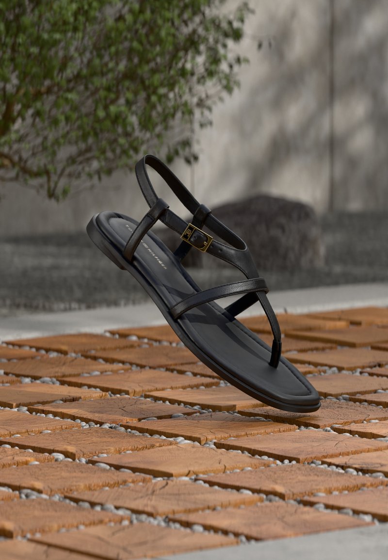 Μαύρο σανδάλι με λουράκια και επίπεδη σόλα, με χρυσή αγκράφα, που αιωρείται πάνω από έδαφος με σχέδιο από τούβλα και βότσαλα, σε εξωτερικό χώρο κοντά σε φυτά.