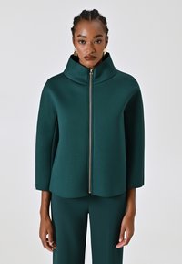 Veste structurée bleu sarcelle avec un col haut, des manches trois-quarts et un détail de fermeture éclair dorée, sur un fond neutre.