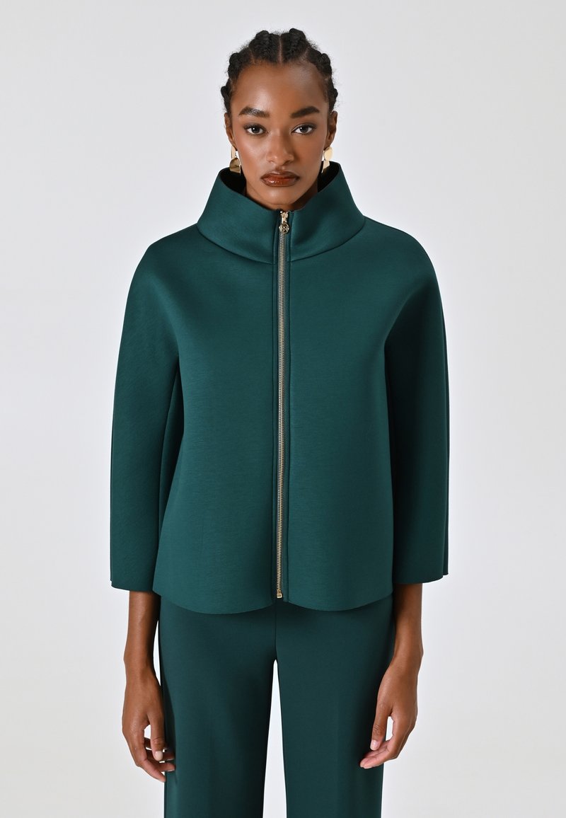 Veste structurée bleu sarcelle avec un col haut, des manches trois-quarts et un détail de fermeture éclair dorée, sur un fond neutre.