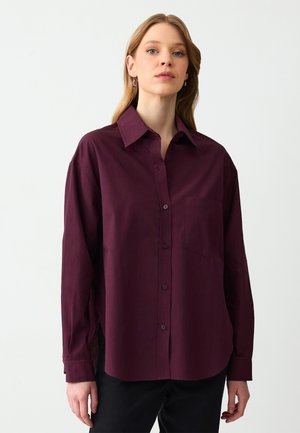 Bordeaux knoopshirt van zachte stof met een kraag, lange mouwen en een borstzak. Bevat zijsplitten en een subtiele textuur.