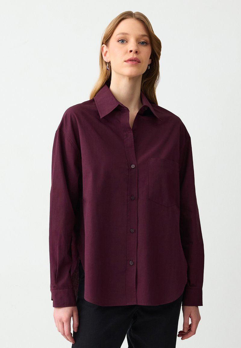 Bordeaux knoopshirt van zachte stof met een kraag, lange mouwen en een borstzak. Bevat zijsplitten en een subtiele textuur.