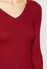 Pull en maille côtelée rouge avec un décolleté en V profond et des manches longues. La texture semble douce, avec un motif de côtes verticales ajoutant du détail.