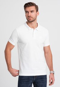 Biała koszulka polo o klasycznym fasonie, krótkie rękawy i zapięcie na guziki pod kołnierzykiem. Posiada subtelne logo i prążkowaną fakturę. Noszona z niebieskimi dżinsami.