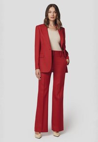 Czerwony, dopasowany garnitur z usztywnianą marynarką i rozszerzanymi spodniami, połączony z beżową bluzką. Materiał wydaje się gładki, z wyraźnymi liniami i minimalnymi detalami.