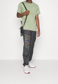 Man som bär en ljusgrön Nike-t-shirt, svarta Nike-byxor med färgglad logotyp, vita sneakers och en liten svart axelremsväska mot vit bakgrund.