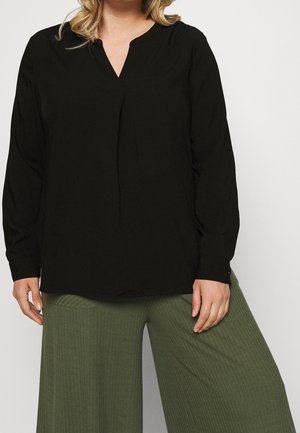 Vrouw die een loszittende zwarte blouse met lange mouwen en een V-hals draagt, met groene geribbelde broek met rechte wijde pijpen, staand tegen een effen achtergrond.