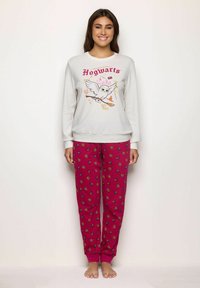 Mujer vestida con sudadera blanca que tiene un búho volador y el texto "Hogwarts", y pantalones rojos con los logotipos "HP" esparcidos, de pie descalza contra una pared blanca.