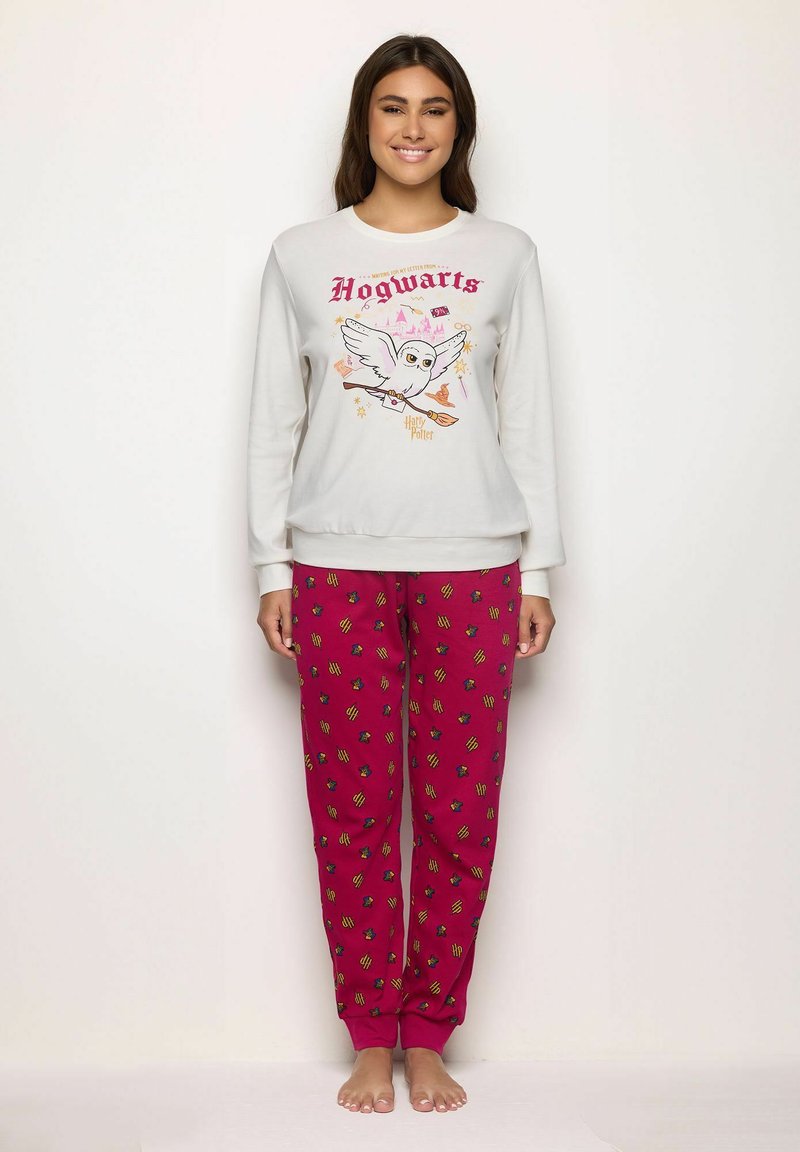 Mujer vestida con sudadera blanca que tiene un búho volador y el texto "Hogwarts", y pantalones rojos con los logotipos "HP" esparcidos, de pie descalza contra una pared blanca.