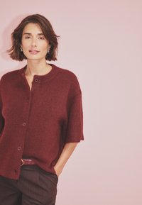 Burgundska oversized kardigan s kratkimi rokavi, teksturiran pleten material, okrogli izrez in zapiranje na gumb, v kombinaciji s temnimi hlačami.