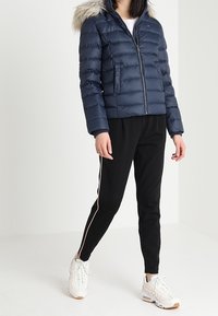 Tmavě modrá péřová bunda s kapucí lemovanou kožešinou, zapínáním na zip a horizontálním prošitím. Nositelná s černými tepláky s růžovým pruhovým detailem na boku.