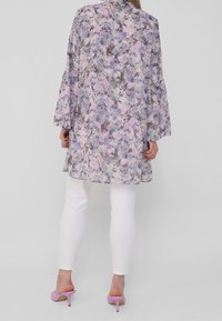 Cardigan léger à imprimé floral dans des tons pastel de violet et de rose, avec des manches larges et une coupe ample, associé à un pantalon blanc et des talons roses.