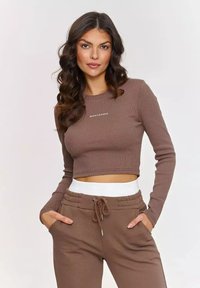 Brązowy prążkowany croptop z długim rękawem z logo "MAKADAMIA", w zestawie z dopasowanymi brązowymi dresami z kieszeniami i białym elastycznym pasem.