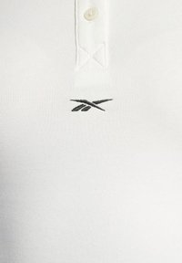 Polo blanc avec deux boutons et un petit logo noir brodé centré sous le col.