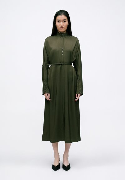 Olive green, long-sleeve dress with a high collar, button front, and belted waist; smooth texture and ankle-length with pointed black shoes.