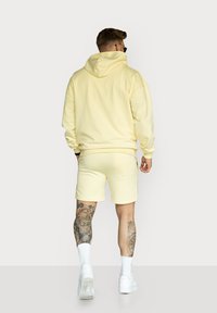 Yellow hoodie and matching shorts, cotton material, relaxed fit, and ribbed cuffs. Notable tattoos visible on legs. White sneakers worn.