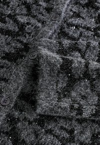 Sivi kardigan s онаčeytuks, s crnim tkanim uzorkom i raspršenim šljokicama. Ima dva tamna dugmeta i detaljno oblikovan ovratnik.