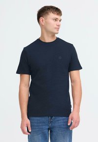 Tamsiai mėlynas trumpomis rankovėmis marškinėliai, pagaminti iš tekstūruotos medžiagos, su apvalia iškirpte ir nedideliu logotipu krūtinėje. Dėvimi su mėlynomis džinsomis.