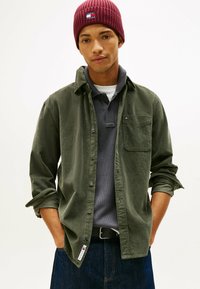 Donkergroene corduroy shirt met een spijkerjack, gedragen over een grijze polo. Draagt een bordeauxrode geribde beanie met een merkpatch op de voorkant.