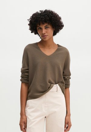 Vrouw met kort krullend haar, een bruine V-hals trui en licht beige hoge taille broek tegen een eenvoudige lichte achtergrond.