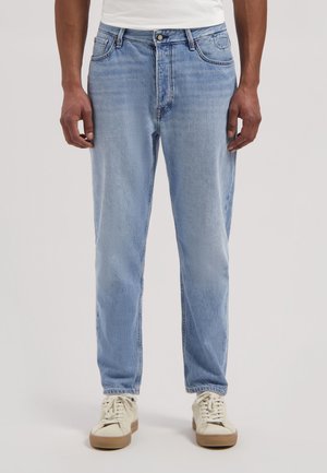 Man die lichtblauwe jeans met rechte pijpen en witte sneakers draagt, staande tegen een effen achtergrond.