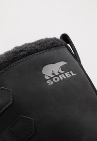Schwarze Lederstiefel mit einem grauen Fleecekragen, Sorel-Logo und dekorativen Nähten sowie einer Zuglasche für einfaches An- und Ausziehen.