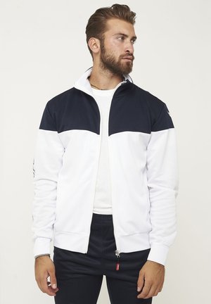 Uomo che indossa una giacca con zip bianca e nera sopra una camicia bianca, che guarda di lato su uno sfondo chiaro e uniforme.