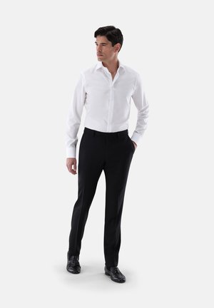 Weißes Hemd mit spitzem Kragen, kombiniert mit schwarzen maßgeschneiderten Hosen. Das Modell trägt schwarze Anzugschuhe und steht vor einem schlichten Hintergrund.