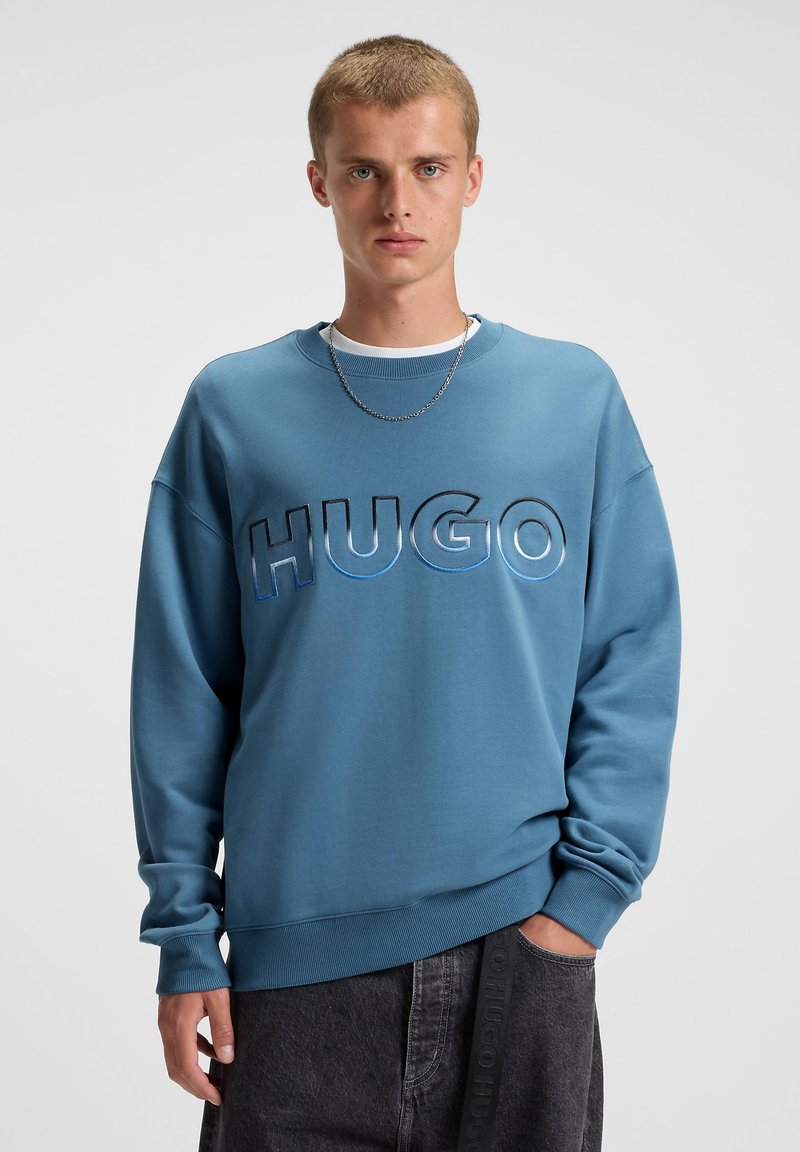 Modrá mikina so zaobleným výstrihom a voľným strihom, s vyšitým logom "HUGO" s gradientovým efektom. Textúrovaná tkanina a rebrované manžety.