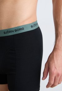Homme portant un boxer noir avec une ceinture verte arborant le nom de marque Björn Borg, se tenant contre un fond clair.