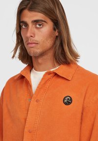 Giacca in corduroy arancione con bottoni a pressione, design con colletto, uno strato interno bianco visibile e una toppa nera sul petto.