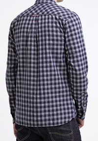 Marineblauwe en grijze geblokte overhem shirt met een kraag, lange mouwen en een gebogen zoom. Bevat subtiele stiksels en een label op de achterkant van de nek.