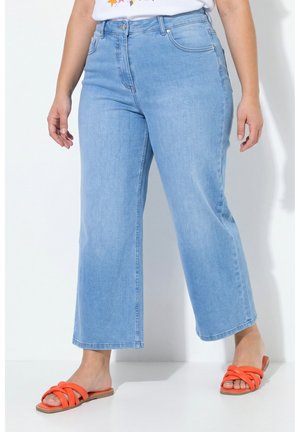 Donna che indossa jeans azzurri a gamba larga e corta e sandali bassi arancioni, in piedi su sfondo bianco.