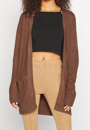Frau trägt eine braune, offene Strickjacke, ein schwarzes, kurzes Oberteil und beige, hoch taillierte Hosen, wobei der Bauchnabel sichtbar ist.