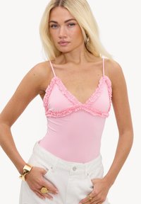 Femme blonde portant un haut sans manches rose avec des bords volantés et un pantalon blanc, accessoirisée avec des bagues en or et un bracelet.