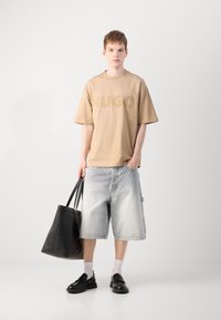 Tricou oversized bej cu imprimeu "HUGO", pantaloni scurți din denim decolorat, geantă din piele neagră tip tote și mocasini negri cu șosete albe.
