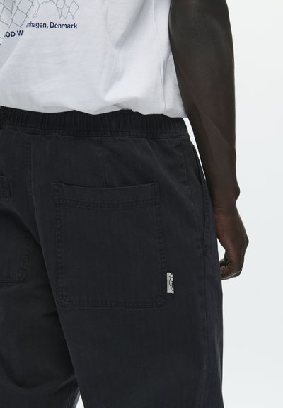 Pantalon noir en coton avec une taille élastique, doté de deux grandes poches arrière et d'une petite étiquette sur le côté. Le tissu a une texture lisse.