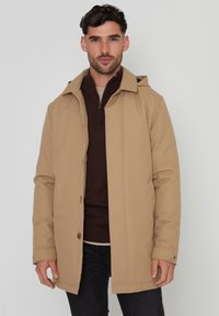 Beige waterdichte jas met een capuchon, knoopaccenten en een rechte snit. Gedragen over een donkerbruin ritsensweater, vergezeld van donkere spijkerbroeken.