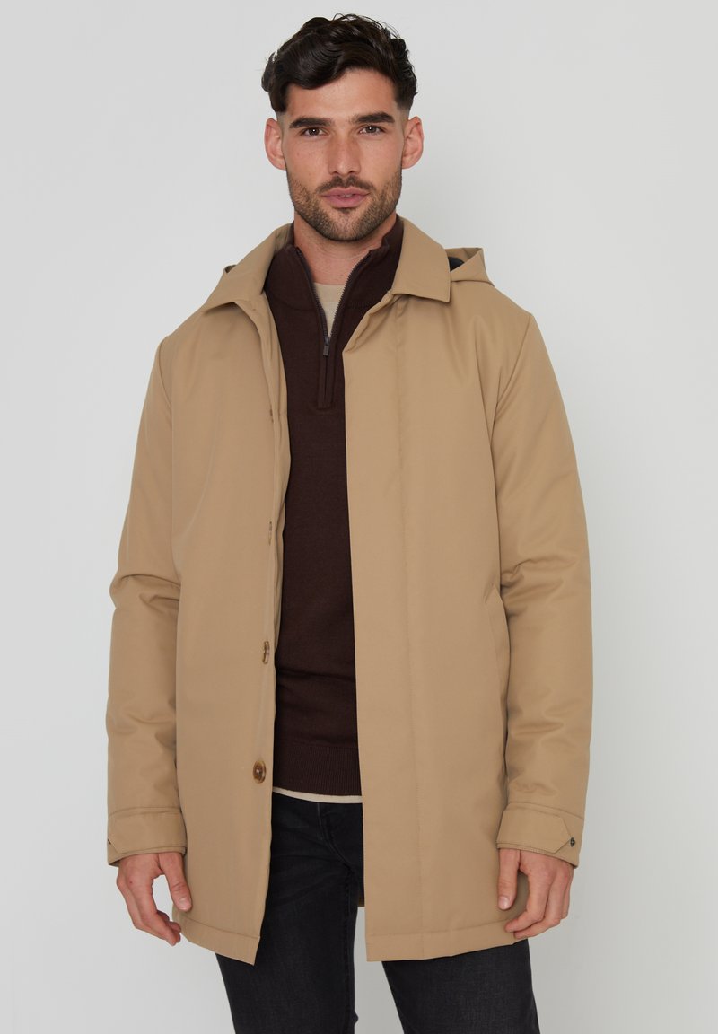 Beige waterdichte jas met een capuchon, knoopaccenten en een rechte snit. Gedragen over een donkerbruin ritsensweater, vergezeld van donkere spijkerbroeken.