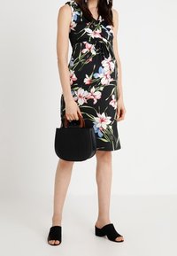 Vestido negro floral con mangas cortas, adornado con flores rosas y azules. Sosteniendo un bolso negro con un asa de madera; lleva mules negras.